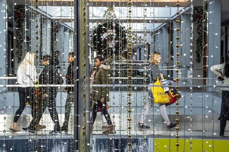 A group of people walking through a decorated shopping mall during the holiday season. The scene is viewed through strings of golden and clear ornaments hanging from the ceiling. One person carries shopping bags, and a large holiday wreath is visible in the background, embodying best practices for festive decor.