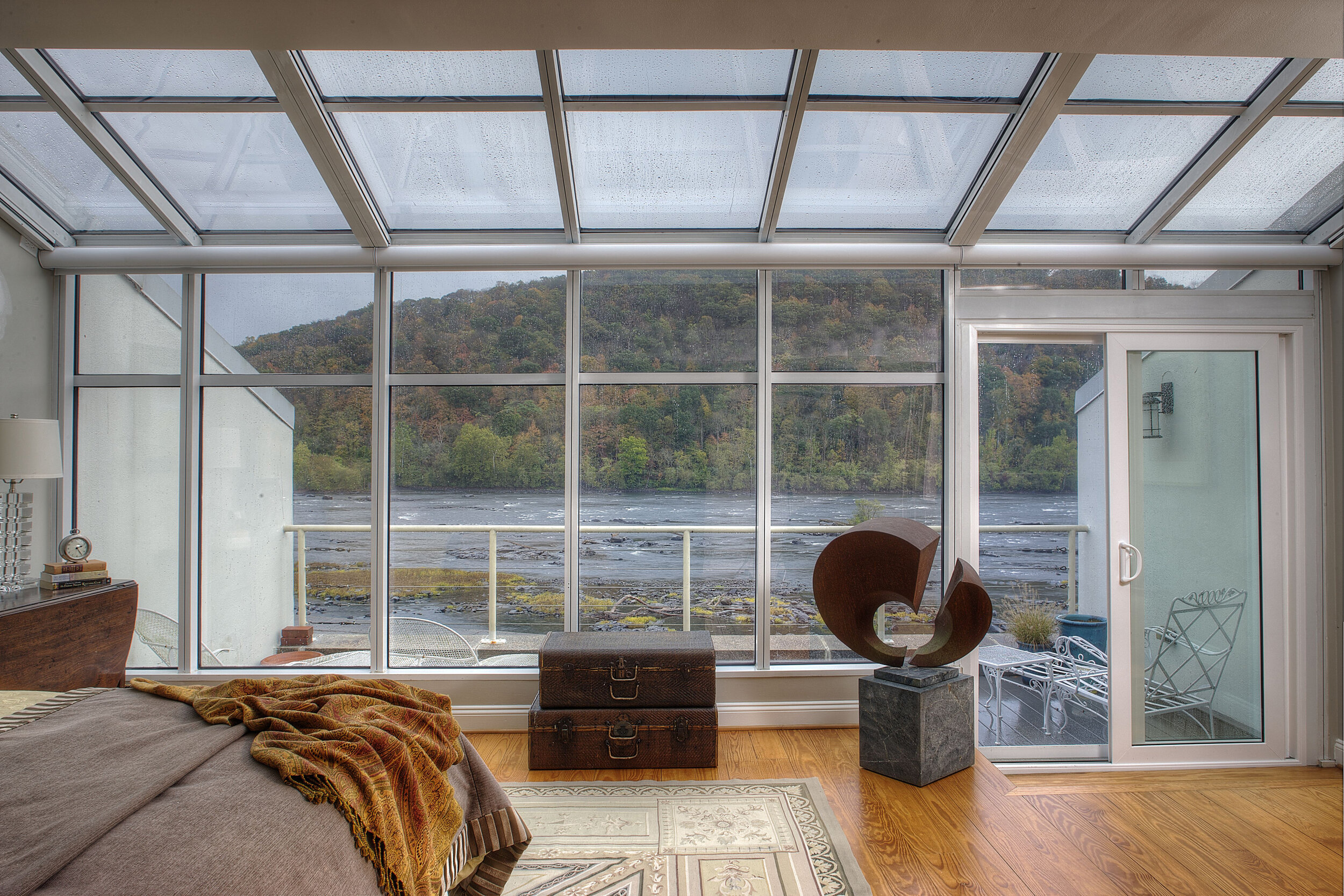 A modern bedroom with large floor-to-ceiling windows offering a scenic view of a river and forested hillside. The room features a bed with a brown blanket, a decorative chest, and a geometric sculpture on a pedestal. Outside the motorized sliding glass door, there is a balcony with patio furniture.