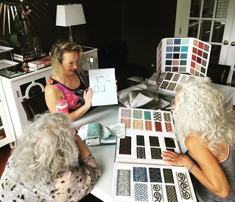 Three women with light hair sit around a table discussing tile samples. One woman, in a dark tank top, holds up a sheet with tile designs while the other two look on. The table is covered with several open sample books showcasing various colorful and patterned tiles, reflecting their meticulous project management skills.