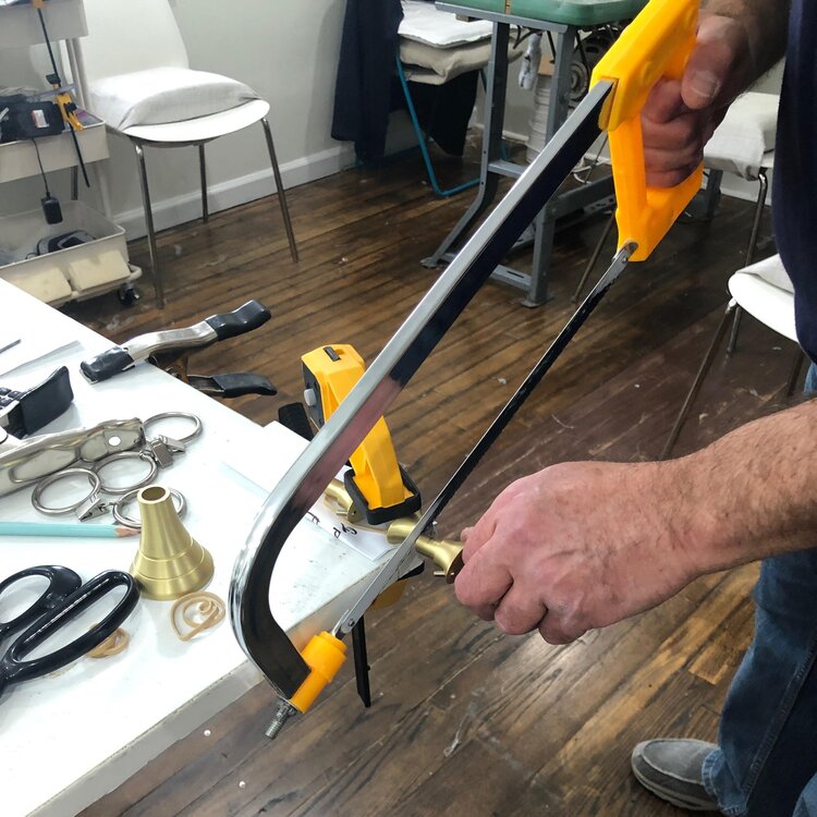 A person uses a hacksaw with a yellow handle to cut a metal piece, showcasing adept problem solving. The workspace, perfect for drapery hardware projects, has wood flooring and a white table cluttered with various tools including scissors, pliers, and a clamp. A white chair and additional equipment are visible in the background.