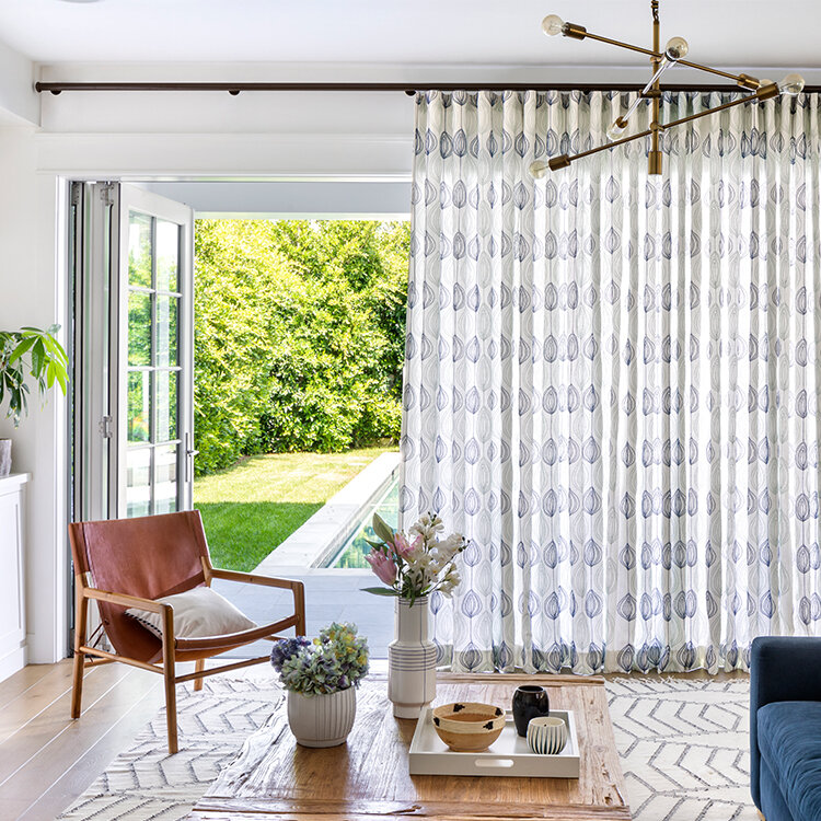 A modern living room with light wood flooring, a brown leather chair, a wooden coffee table, and a blue couch partially visible. The room features large, patterned curtains and folding glass doors opening to a lush green garden with a narrow pool. Discover how to decorate doors like the pros with these inspiring ideas! A brass chandelier hangs from the ceiling.