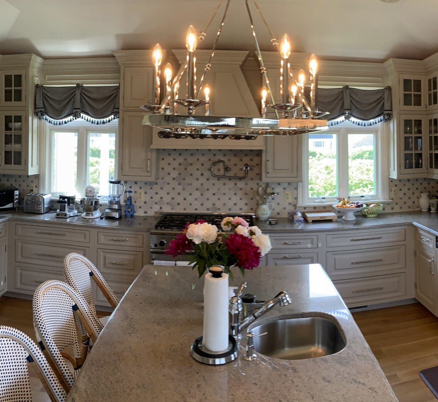 A spacious, modern kitchen with cream-colored cabinetry and granite countertops. A chandelier hangs over a central island with a sink and vase of flowers. Stainless steel appliances gleam, while large windows dressed with decorative curtains showcase a scenic view. Two wooden stools sit by the island.