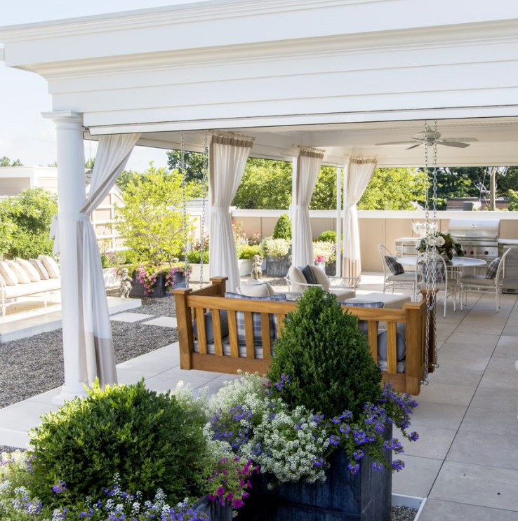 A spacious rooftop patio with a white pergola and curtains exemplifies comfort. It features cushioned seating, a wooden coffee table, and an outdoor dining area with white metal chairs and tables. Adorned with potted plants, flowers, boxwood shrubs, and patio shades, it offers views of nearby greenery.