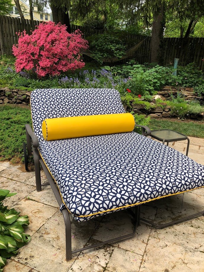 A patio lounger with a patterned navy and white cushion and a yellow bolster pillow sits on a stone patio. The lounger, perfect for outdoor living, faces a lush garden featuring dense greenery and vibrant pink blooming flowers. A small glass-top side table is positioned next to the lounger, elevating the space.