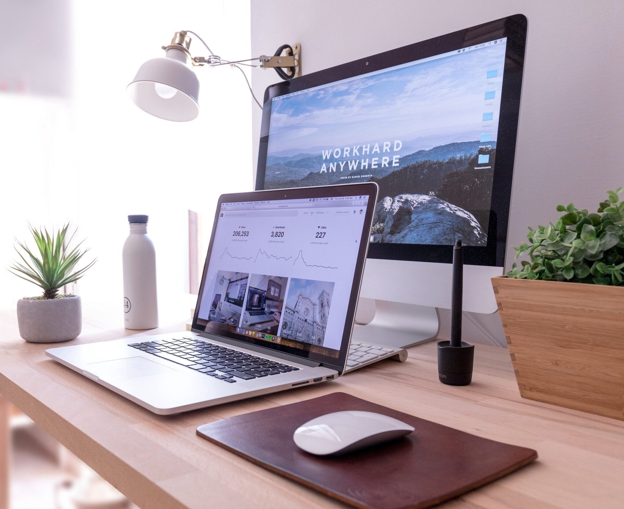 A well-organized workspace featuring a wooden desk with a potted plant, a water bottle, a notepad, a mouse on a brown mouse pad, a pen holder with a stylus, and a MacBook displaying charts from the project management system. A desktop monitor shows the "Work Hard Anywhere" wallpaper under a wall-mounted lamp.