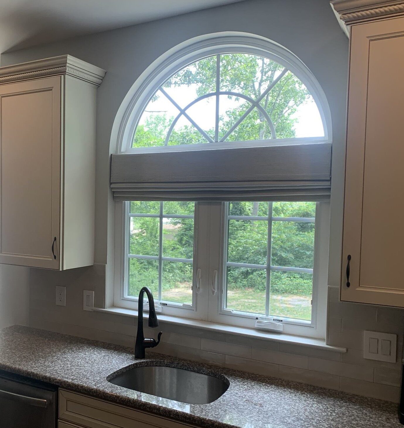 A kitchen with a granite countertop, stainless steel double sink, and black faucet. Above the sink is an arched window with a view of trees, complemented by a smaller rectangular transom window. Light-colored cabinets surround the area, and a sliding glass door with vertical blinds is visible to the left.