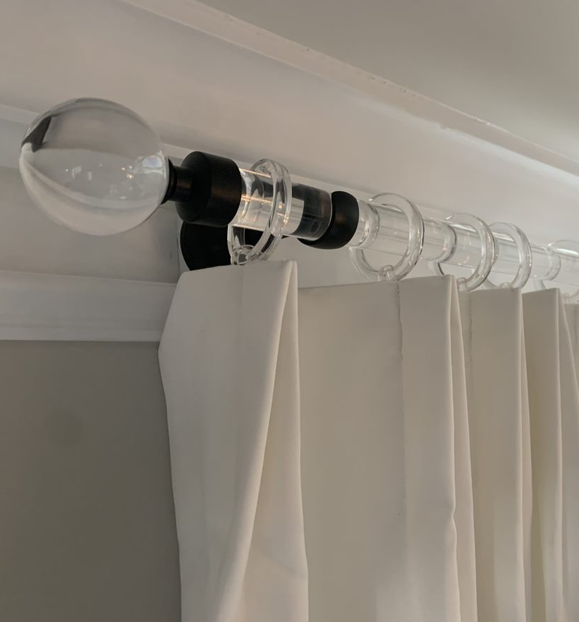 A clear acrylic curtain rod with a spherical finial at the end is mounted near the ceiling. White curtains hang from the rod using transparent hooks. The black brackets are attached to a white wall, providing a minimalist and modern look—perfect drapery tips for using elegant acrylic hardware.