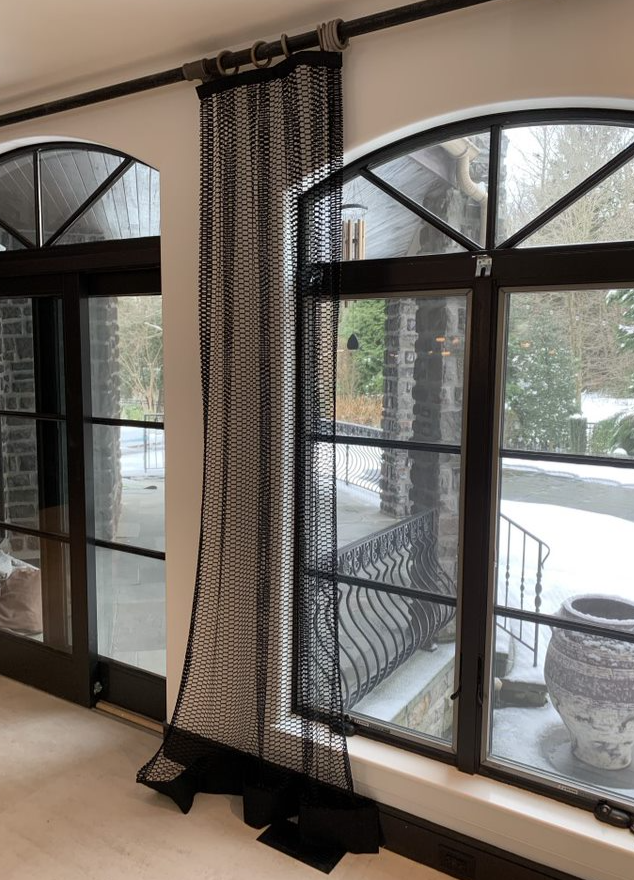 A room with large arched windows featuring black metal frames and hardware. Outside, a snow-covered patio and landscape are visible. Inside, a black mesh curtain—often considered challenging fabrics—hangs from a rod running across the left window, its length pooling slightly on the floor.