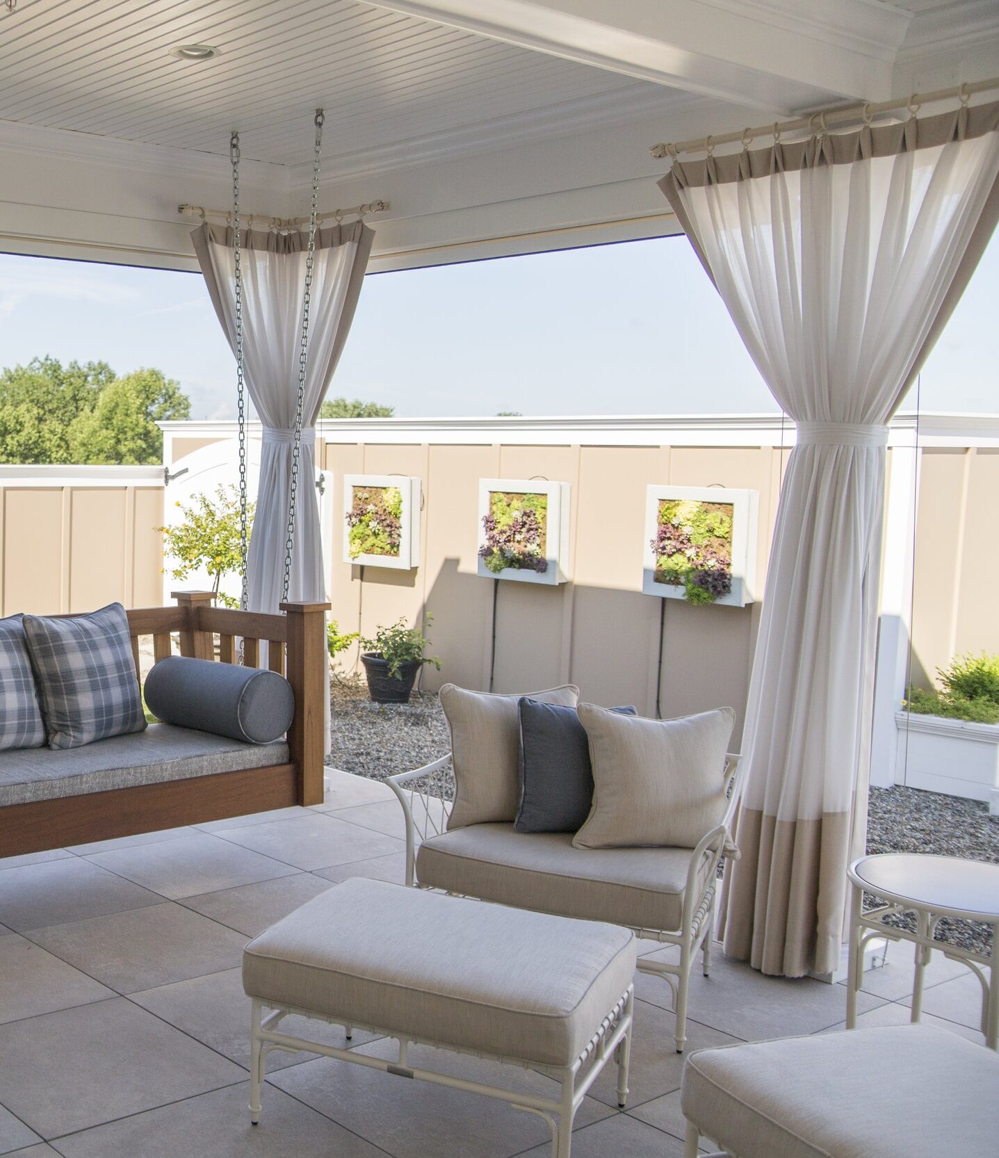 A cozy patio features a suspended wooden bench with gray plaid cushions and round bolster pillows. White metal chairs and ottomans, also with beige cushions, surround the area. Beige and white curtains hang from posts, providing the perfect patio shades. A planter with greenery sits in the corner by a beige fence adorned with small, framed greenery sections.