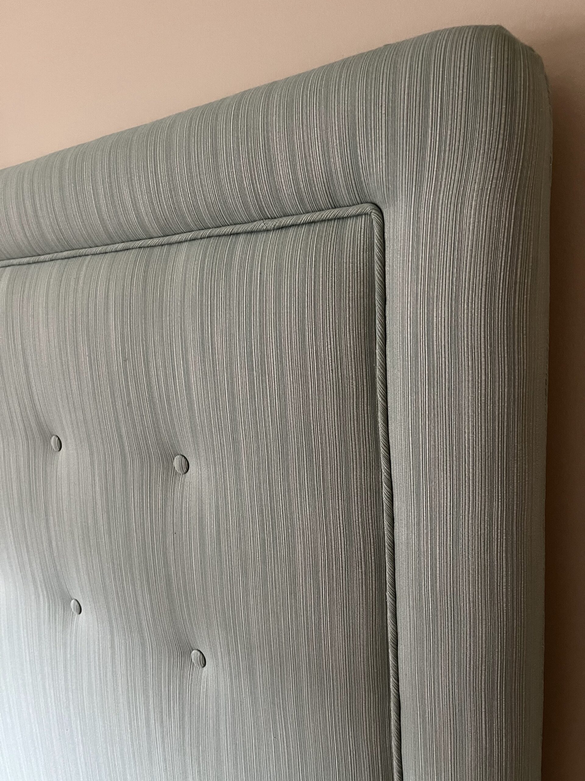 The image shows a close-up of a tall, padded headboard of a bed. The headboard, part of our custom beds collection, is upholstered in light green fabric with a subtle linear texture. It has a soft, plush appearance and features a slightly rounded top edge with evenly spaced buttons creating a tufted effect.