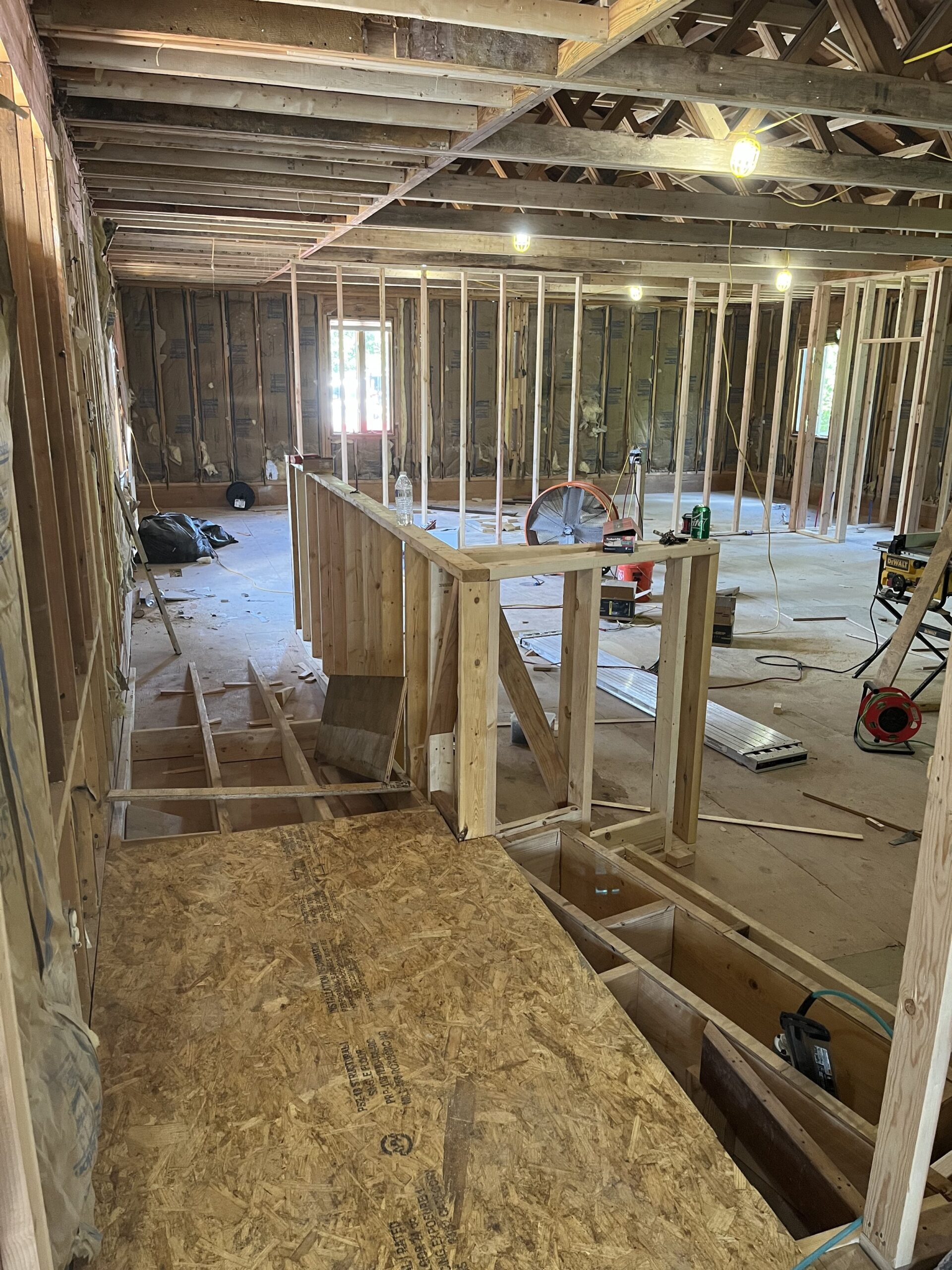 A partially constructed building interior at 67 Almshouse reveals exposed wooden framing and ceiling beams. The plywood flooring and visible insulation in the walls promise a future home. Tools, ladders, and construction materials are scattered throughout, while bright working lights hang from the ceiling beams.