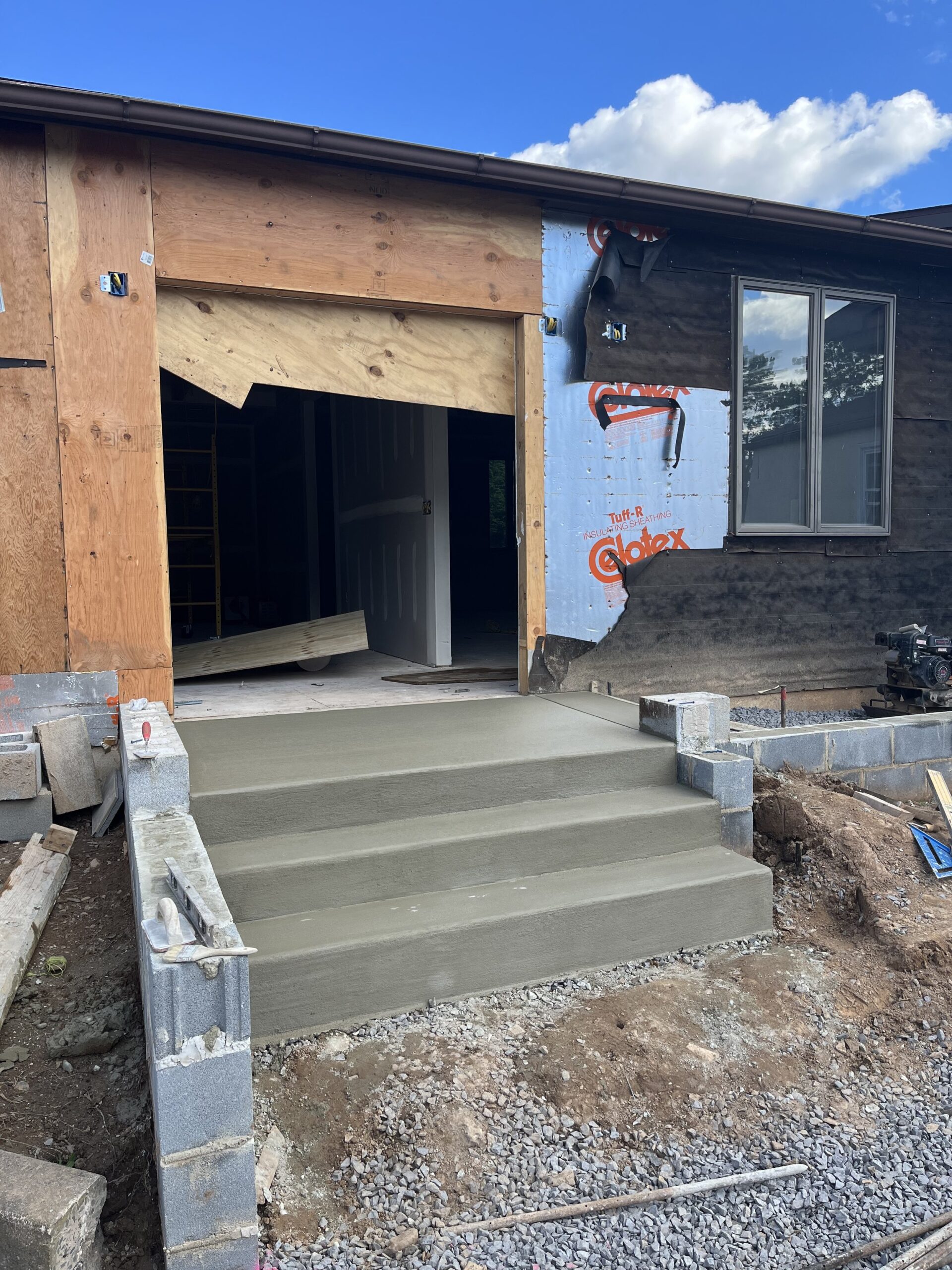 At 67 Almshouse, a house under construction features a newly poured concrete front staircase leading to a doorway in the unfinished facade. Wooden and plastic barriers surround the area, and construction materials like cinder blocks, wooden boards, and gravel are scattered around the site.