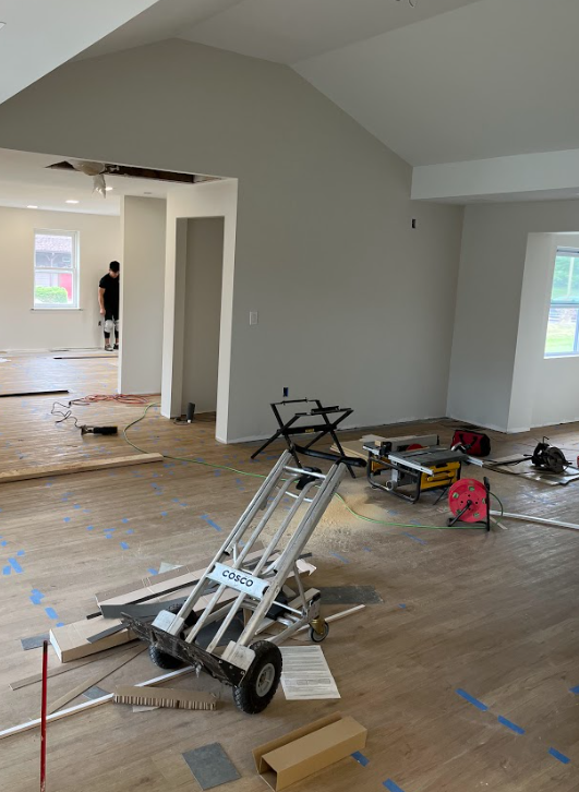 A room at 67 Almshouse is under renovation with gray walls and wood flooring. Construction materials and tools, including a dolly, saw, and power cord, are scattered across the floor. Two windows allow natural light in as one person is visible in the background working on the flooring, nearing the home stretch.