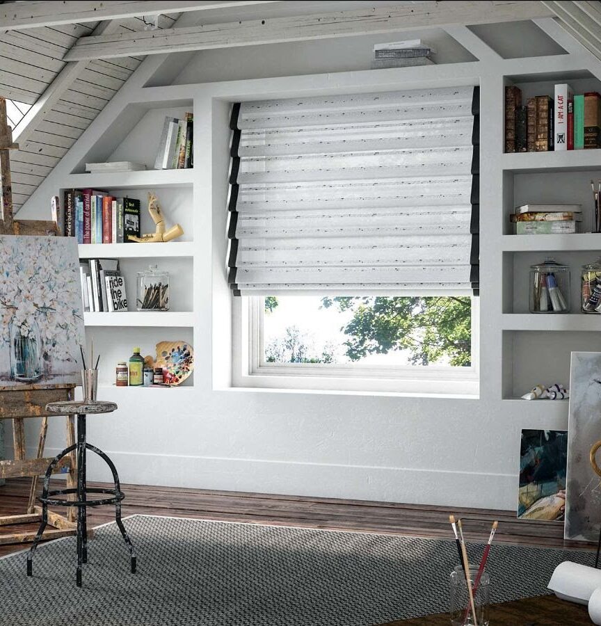 A cozy artist's studio with skylights and a window that overlooks greenery. Shelves on both sides of the window hold books, jars of paintbrushes, and art supplies. Woven blinds add charm to the rustic wooden floor space, where an easel with a floral painting sits beside a stool, various artworks leaned against the wall.