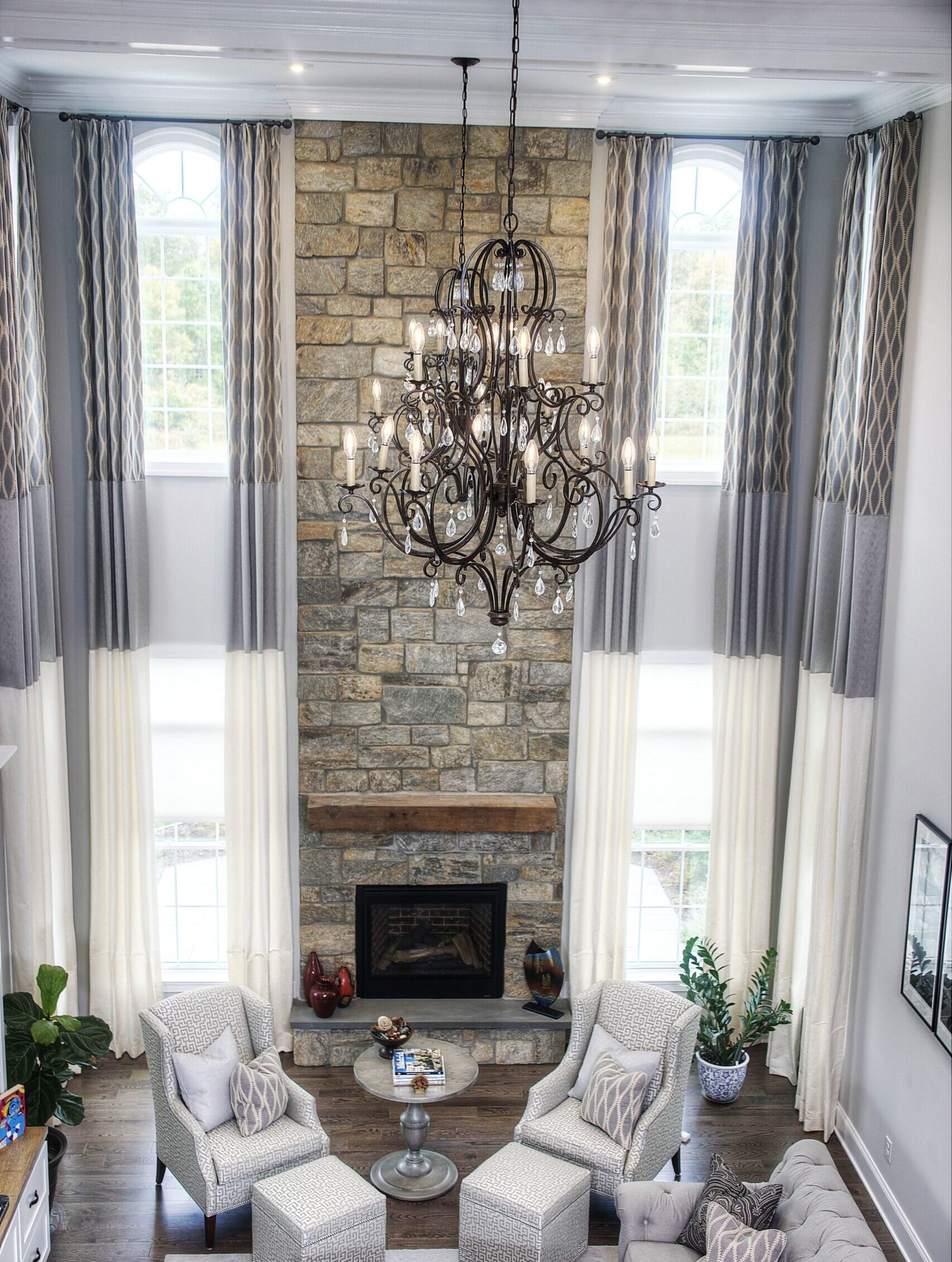 A spacious living room features high ceilings with dressing two-story windows adorned with gray and white curtains. A large, ornate chandelier hangs from the ceiling. The focal point is a stone fireplace with a wooden mantel. The room includes cozy seating, a gray sectional sofa, and a patterned rug.