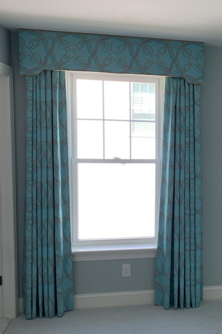 A spacious room with light blue walls and carpet. Two large windows are adorned with blue curtains and matching valances featuring a gold swirl pattern. Under the right window, there is a small wooden table with two drawers. A door is partially visible to the left, adding to the room's charm.