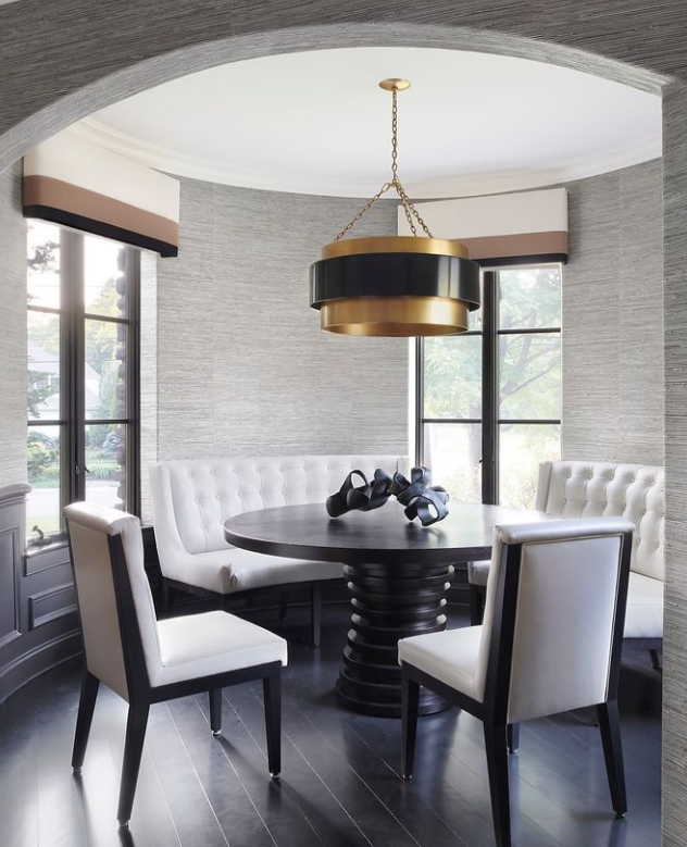 A chic dining room nook features a round black table with a unique spiral base, surrounded by white upholstered chairs with black frames. A large, modern black and gold pendant light hangs above. The room has grey textured walls, window treatments complementing large windows with light pouring in, and dark hardwood floors.