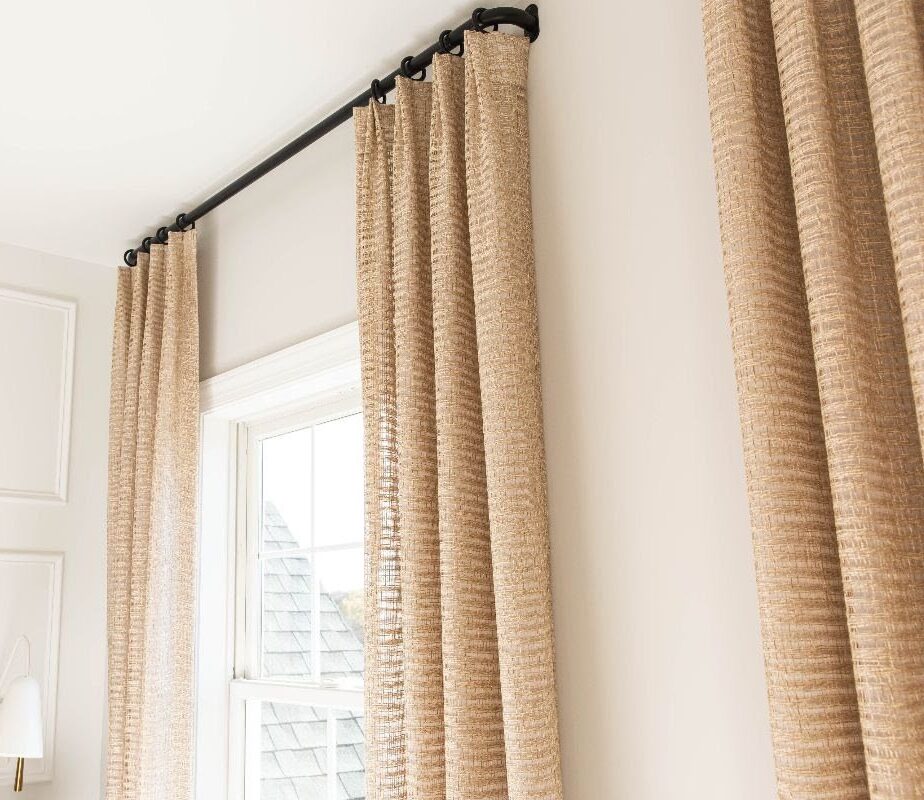 The image shows beige curtains hanging from a black curtain rod above a window in a bright room. The window is partially visible and offers a view of a nearby building's roof. A wall-mounted lamp and the corner of a gray upholstered headboard are seen on the left side, indicating no fabric issues such as textile instability.