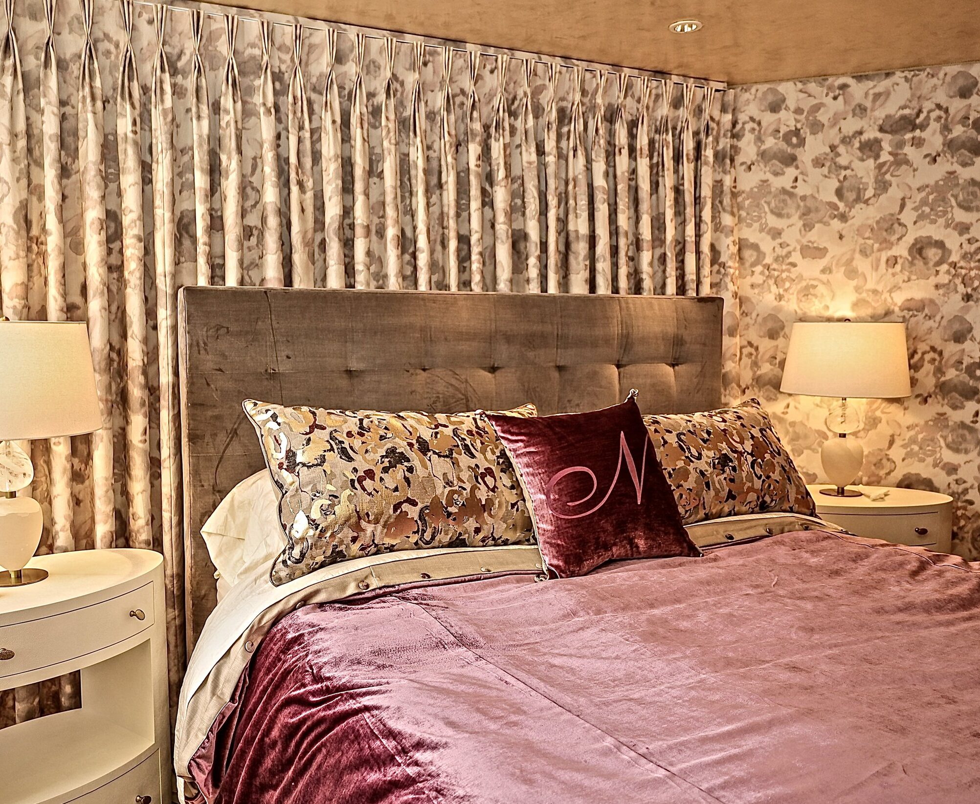 A cozy bedroom features a tufted gray headboard against floral patterned, upholstered walls. The bed is adorned with a velvety mauve bedspread and multiple pillows, including a central maroon one with a monogram. Two white nightstands with lamps are on either side of the bed, and window treatments match the wallpaper.