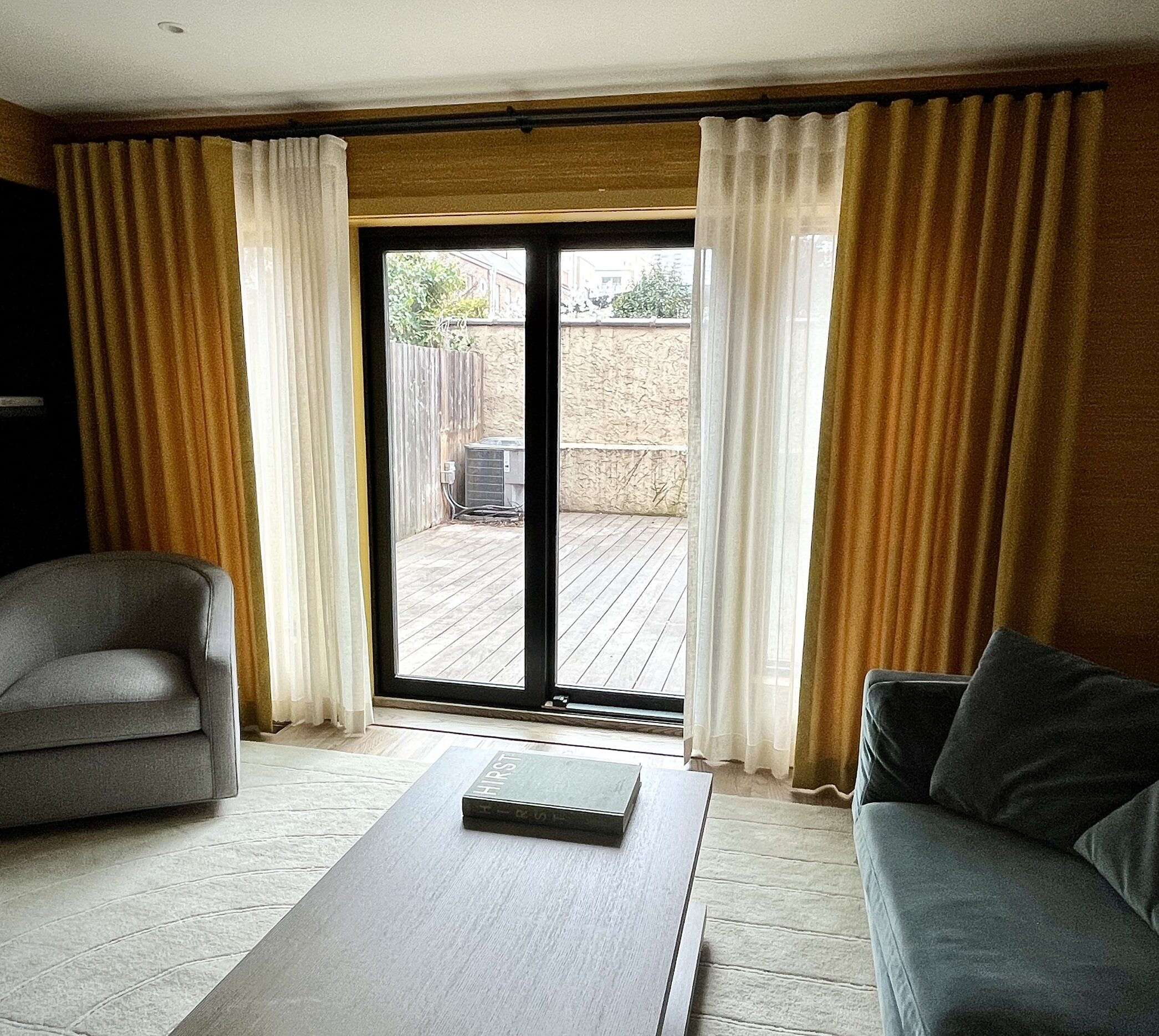 A cozy, modern living room featuring a grey armchair on the left, a dark grey sofa on the right, and a light-colored wooden coffee table in the center. The room has large glass sliding doors with H-Rail Hardware and light brown and white curtains, opening to a wooden deck outside. Minimalist décor and a neutral color palette define the space.