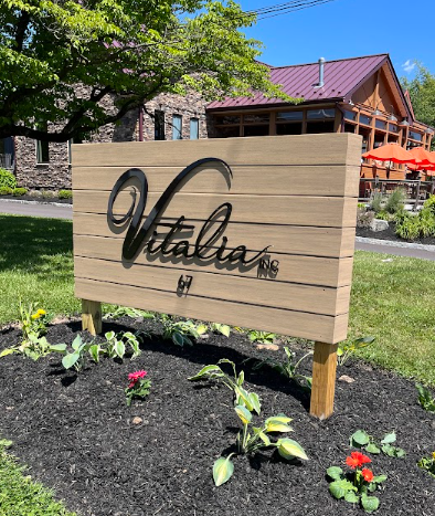 A wooden sign with "Vitalita" engraved on it stands on two wooden posts in a mulched bed with small plants and flowers. The bed is surrounded by grass at 67 Almshouse. In the background, there's a building with a stone facade, a metal roof, and outdoor seating with orange umbrellas. Trees and a clear blue sky are also visible.