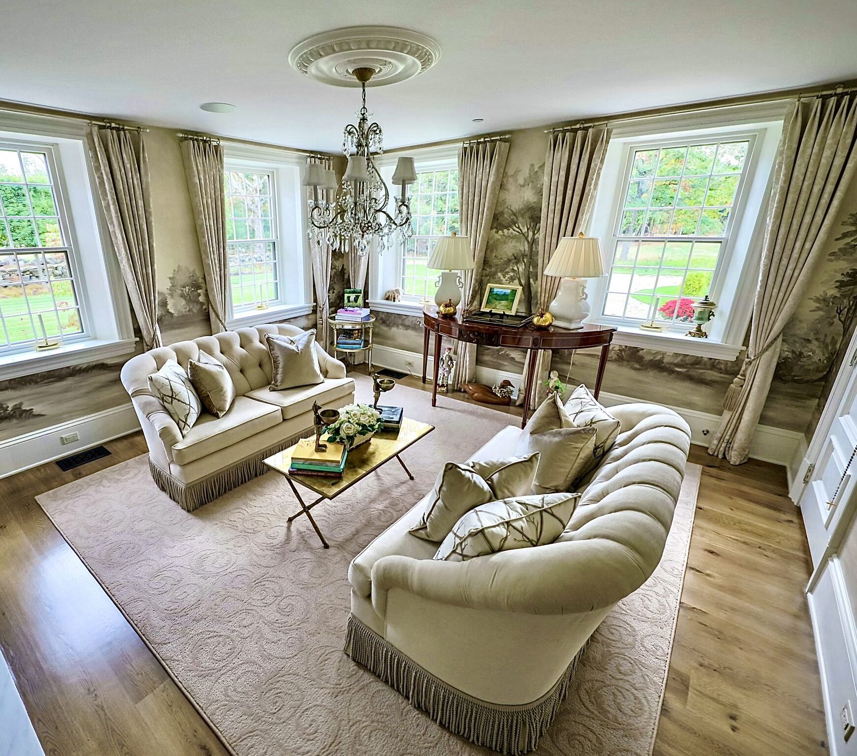 A spacious living room in the New Hampshire home, with elegant decor featuring two cream-colored sofas facing each other with a glass-top coffee table between them. The room has large windows with draped curtains, a chandelier, and a mural wallpaper. It’s part of Phase 2 of our interior design project.