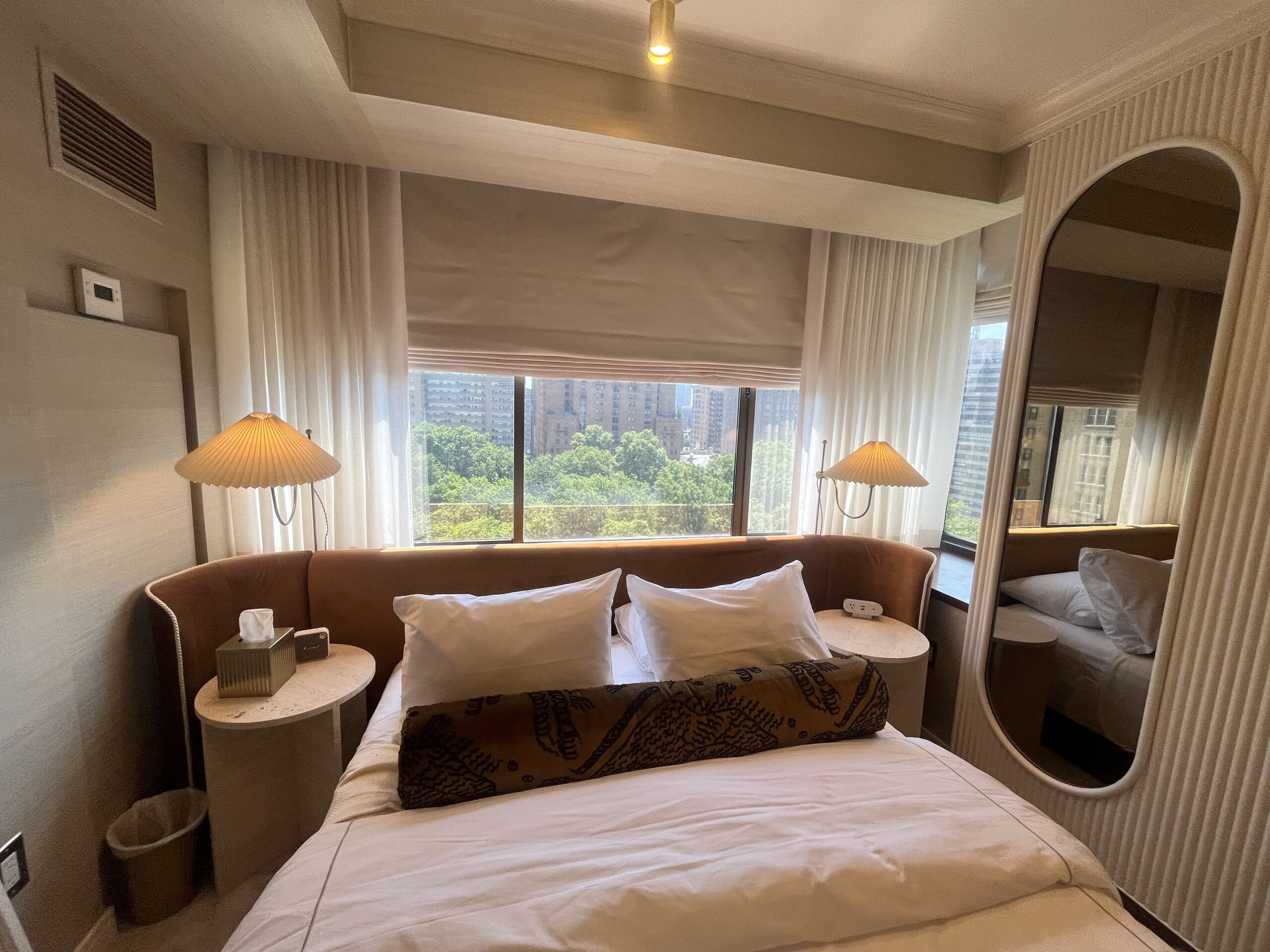 A cozy hotel room with a large bed, white linens, and brown velvet headboard. Custom window treatments frame large windows that reveal trees and tall city buildings, enhancing the luxury interior design of this inviting space.