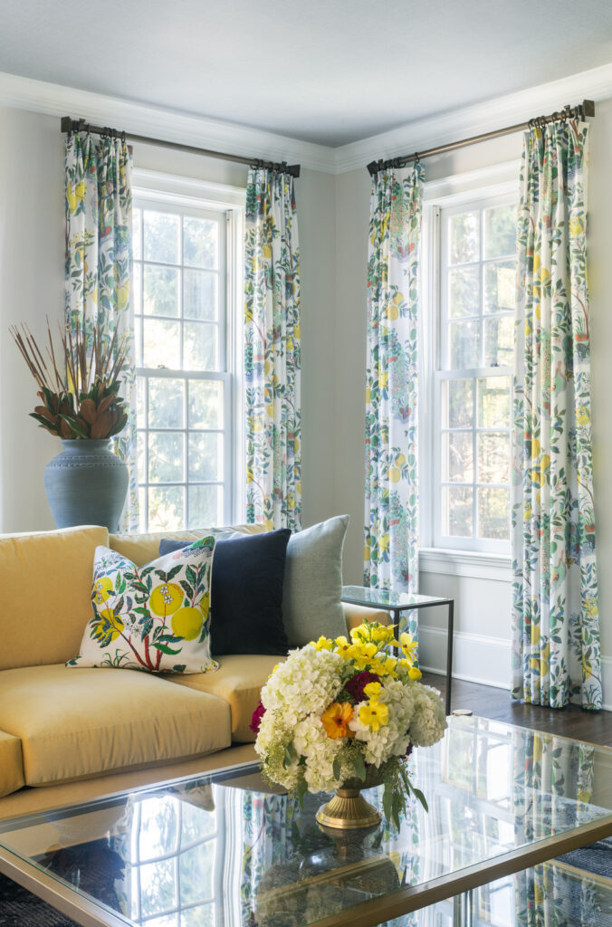 Bright living room with two tall windows dressed in floral curtains, showcasing the style of whole-home window treatments by Liz Walton Home. A yellow sofa features colorful cushions, one with a floral print. A glass coffee table displays a vase of white and yellow flowers, while a large blue vase with branches sits in the corner.
