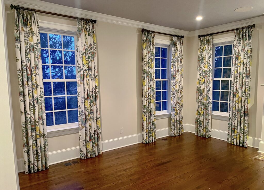 Three tall windows with white trim are covered by floor-length, floral-patterned curtains featuring green, yellow, and gray accents—an elegant example of thoughtful window treatments. The room has beige walls, hardwood floors, and white crown molding, with trees visible through the windows at dusk.