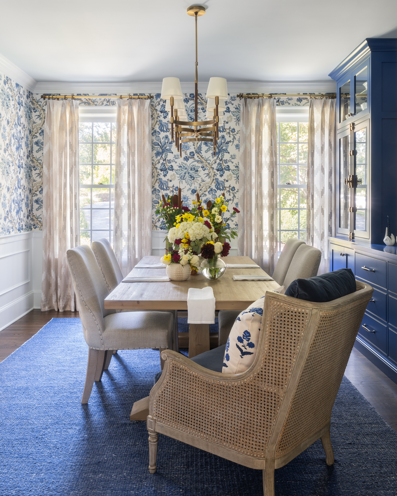 Bright dining room with a wooden table, six light upholstered chairs, floral centerpiece, blue floral wallpaper, sheer window treatments, blue carpet, and a blue china cabinet. Natural light streams through three tall windows behind the table and chairs—an inviting Liz Walton Home design perfect for whole-home gatherings.