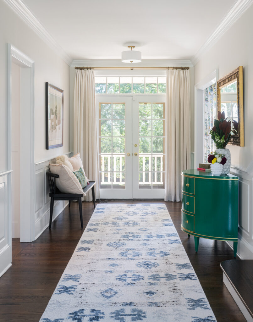 Bright hallway with dark wood floors, white walls, a blue-patterned rug, a green chest with decor and books, a black bench with pillows, cream curtains from Liz Walton Home, and large windows and French doors letting in natural light—showcasing the beauty of thoughtfully selected whole-home window treatments.