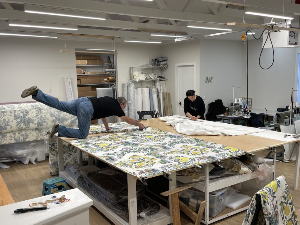 Two people work in a fabric workshop. One person kneels on a table, stretching to measure floral fabric, while another sits, sewing. The bright room, filled with shelves of fabric rolls, tools, and sewing machines, suggests the creative hub where window treatments for spaces like those designed by Liz Walton Home might begin their journey.