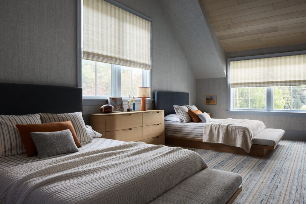 Two modern twin beds with gray headboards and neutral bedding sit in a spacious, light-filled bedroom at the Flacco Family Home. A wooden dresser separates the beds, while large windows with light blinds line the gray walls, giving full scope to natural daylight. A striped rug covers the floor, enhancing the room’s cozy, minimalist feel within this thoughtfully designed building.