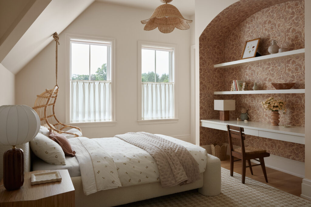 Cozy bedroom with a bed, neutral bedding, and a hanging rattan chair by two bright windows. Built-in desk with shelves, decorated with vases, books, and art, is set in an alcove with patterned wallpaper. Light wood and soft textures create a warm, inviting atmosphere—perfectly reflecting the thoughtful building scope of the Flacco Family Home.