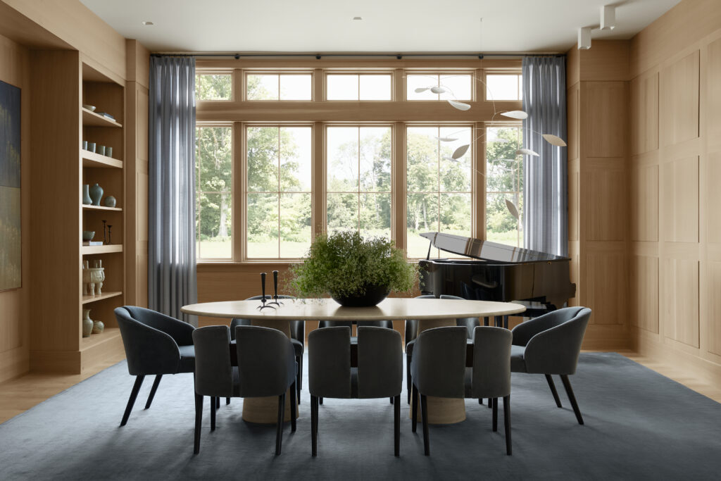 A modern dining room in the Flacco Family Home features a large oval wood table, gray upholstered chairs, a black grand piano, tall curtained windows, built-in shelving, wood panel walls, and a blue rug. A plant centerpiece and minimalist chandelier complete the elegant, airy space—offering a glimpse into the full scope of thoughtful home building throughout.