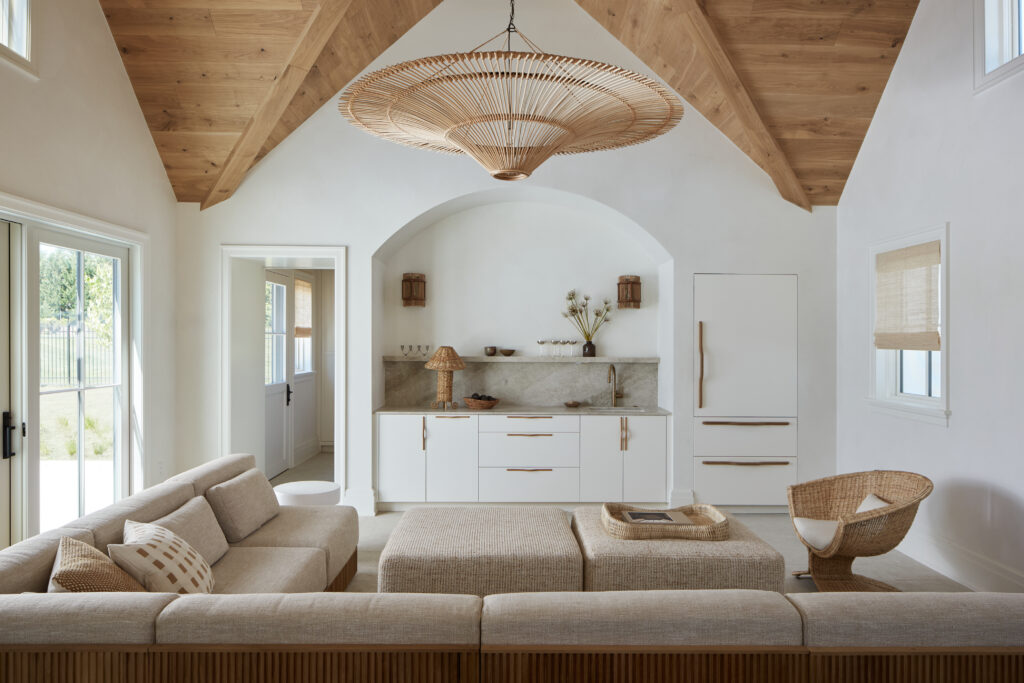 Bright, airy living room with vaulted wood ceiling, rattan chandelier, beige sectional sofa, woven chair, neutral décor, and minimalist white kitchen with marble backsplash. Large windows let in natural light. The full scope of décor feels coastal and serene—perfect for a welcoming Flacco Family Home.