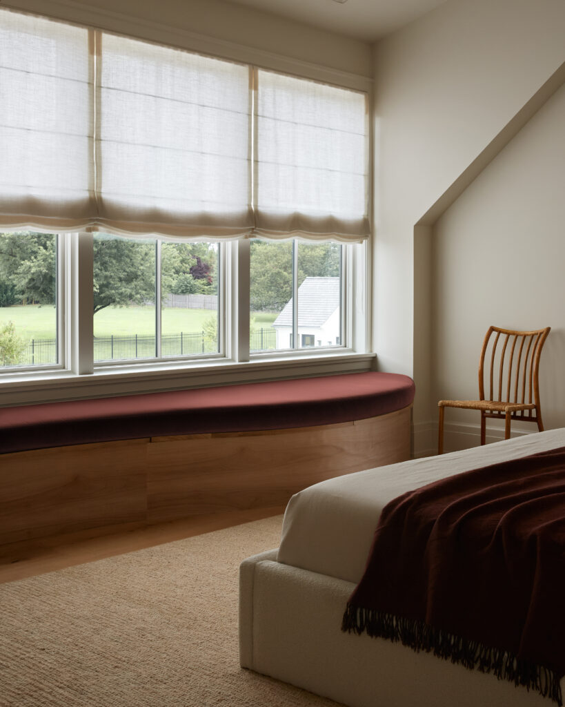 A bedroom in the Flacco Family Home features a bed covered in a dark blanket, a wooden chair, a built-in wooden window seat with a maroon cushion, beige carpet, and large windows with sheer white Roman shades overlooking a green yard and trees—a serene retreat thoughtfully designed within the home’s detailed building scope.