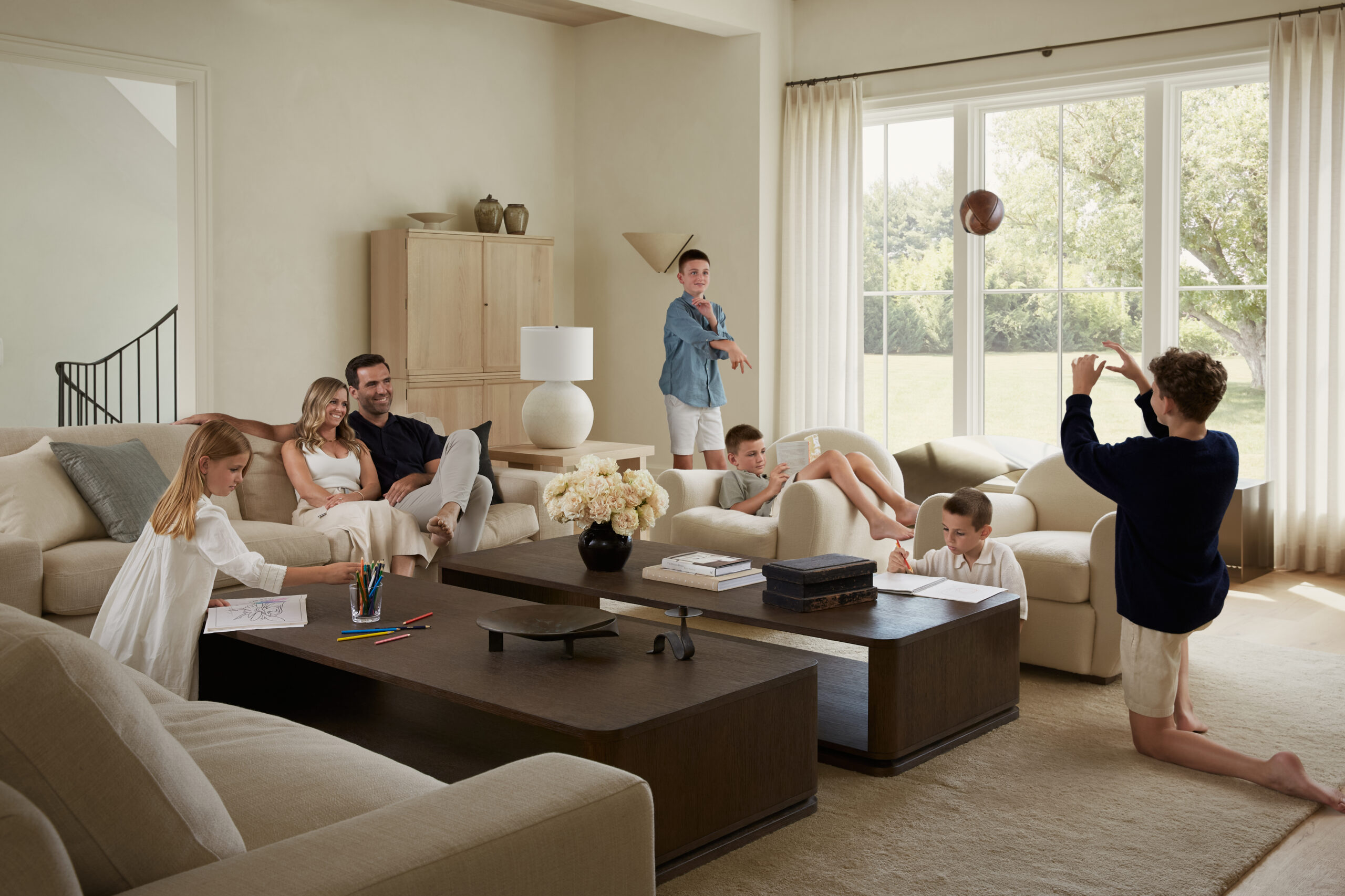 A family sits in a bright living room with beige sofas in the Flacco Family Home. Two adults relax on a couch, surrounded by four children: one drawing, one kneeling and tossing a ball, one on a tablet, and one sitting on the sofa. Large windows—part of the thoughtful home construction—show a sunlit yard outside.