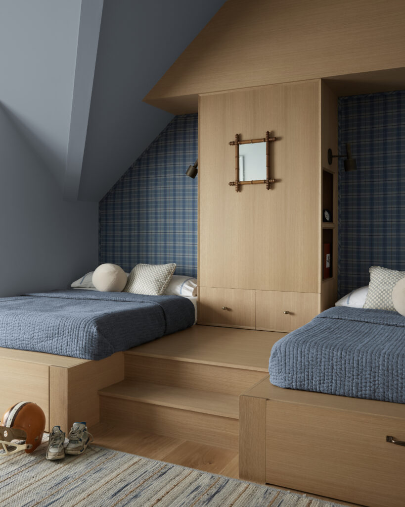 Two twin beds with light wood frames and blue bedding sit on either side of built-in drawers and shelves in the Flacco Family Home. The walls are blue plaid and solid blue, adding charm to the carefully considered building scope of the room. A helmet, shoes, rug, and pillows are visible, while steps lead up to the beds. Behind the curtain, a nautical mirror hangs between them, adding a coastal touch.